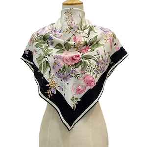 Pode incluir: Um lenço quadrado com centro branco e um padrão floral de rosas cor-de-rosa, flores roxas e folhas verdes. O lenço tem uma borda preta larga com uma risca branca fina e o texto Yves Saint Laurent impresso no canto inferior esquerdo.
