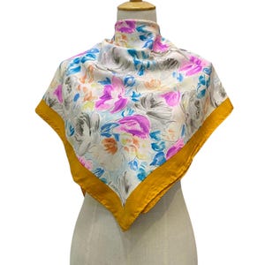 May include: A square silk scarf featuring a floral design in pink, blue, and yellow hues against a cream backdrop. It has a gold border and is displayed on a mannequin. The word "Longhi" is visible.