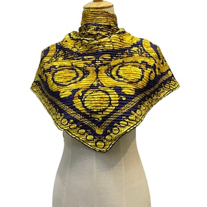 Puede incluir: Un pañuelo cuadrado plisado con una base de color azul marino profundo y un vibrante patrón ornamental en tono dorado. El diseño incluye intrincados motivos circulares, filigranas en espiral y un borde decorativo, todo en amarillo metálico.