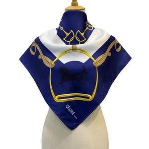 Puede incluir: Un pañuelo cuadrado de seda en azul marino y blanco, con un diseño dorado de carruaje y caballo. El pañuelo tiene un borde dorado y la palabra "CELINE" está impresa en blanco. El pañuelo se exhibe en un maniquí.