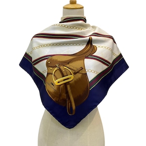 May include: A square silk scarf with a brown equestrian saddle design and gold hardware. The scarf has a white background with gold chain and red and green stripe accents, framed by a navy blue border. The brand name "LONGCHAMP" is visible.
