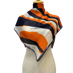May include: A white, orange and navy blue silk scarf with an abstract pattern. The scarf has a purple and white label that reads "Christian Dior Paris".