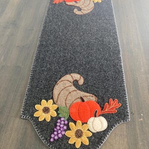 May include: A gray herringbone-patterned table runner with a white stitched edge. The runner features appliqued fall harvest motifs, including a cornucopia, pumpkins, sunflowers, grapes, and fall leaves. The design is symmetrical, with elements at both ends.