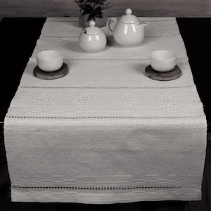 Op de afbeelding: Een witte gehaakte tafelloper met een delicate kantversiering en een herhalend patroon van kleine cirkels. De loper ligt op een zwarte tafel met witte keramische theekopjes en een theepot.