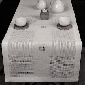 Op de afbeelding: Een witte linnen tafelrunner met een geometrisch patroon. De runner heeft een subtiel rasterontwerp met vierkante vormen die in de stof zijn geweven. De runner ligt op een zwart oppervlak met twee witte kommen en een theepot.