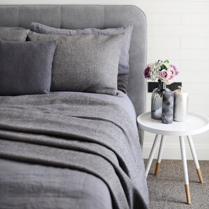 Könnte beinhalten: Ein graues Bett mit passendem Kopfteil, Kissen und Decke. Ein weißer Beistelltisch beherbergt eine Vase mit Blumen, eine Kerze und eine dekorative Flasche. Der Raum hat eine neutrale Farbpalette.