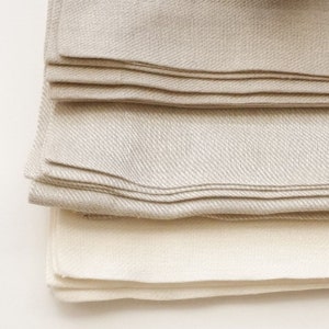 Könnte beinhalten: Ein Stapel aus drei Leinen-Servietten in Beige und Weiß. Die Servietten sind gefaltet und haben ein dezentes Fischgrätenmuster.