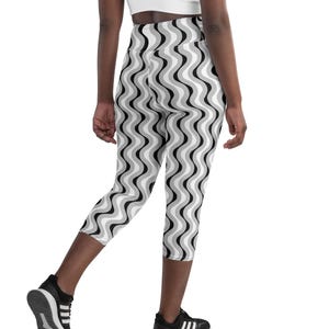 May include: Capri leggings featuring a high-waisted design and a repeating vertical wavy pattern in shades of black, white, and grey. The leggings extend to mid-calf length and are paired with black and white athletic trainers.