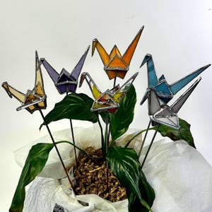 May include: A collection of five colorful stained glass origami crane garden stakes. The cranes are in shades of yellow, purple, orange, blue, and gray. Each crane is mounted on a black metal stake and placed in a potted plant.