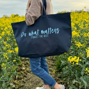Pode incluir: Uma bolsa tote preta com o texto "Do what matters, forget the rest" em letras azuis claras. A bolsa est&aacute; sendo segurada por uma pessoa que est&aacute; vestindo jeans azul e um su&eacute;ter marrom.