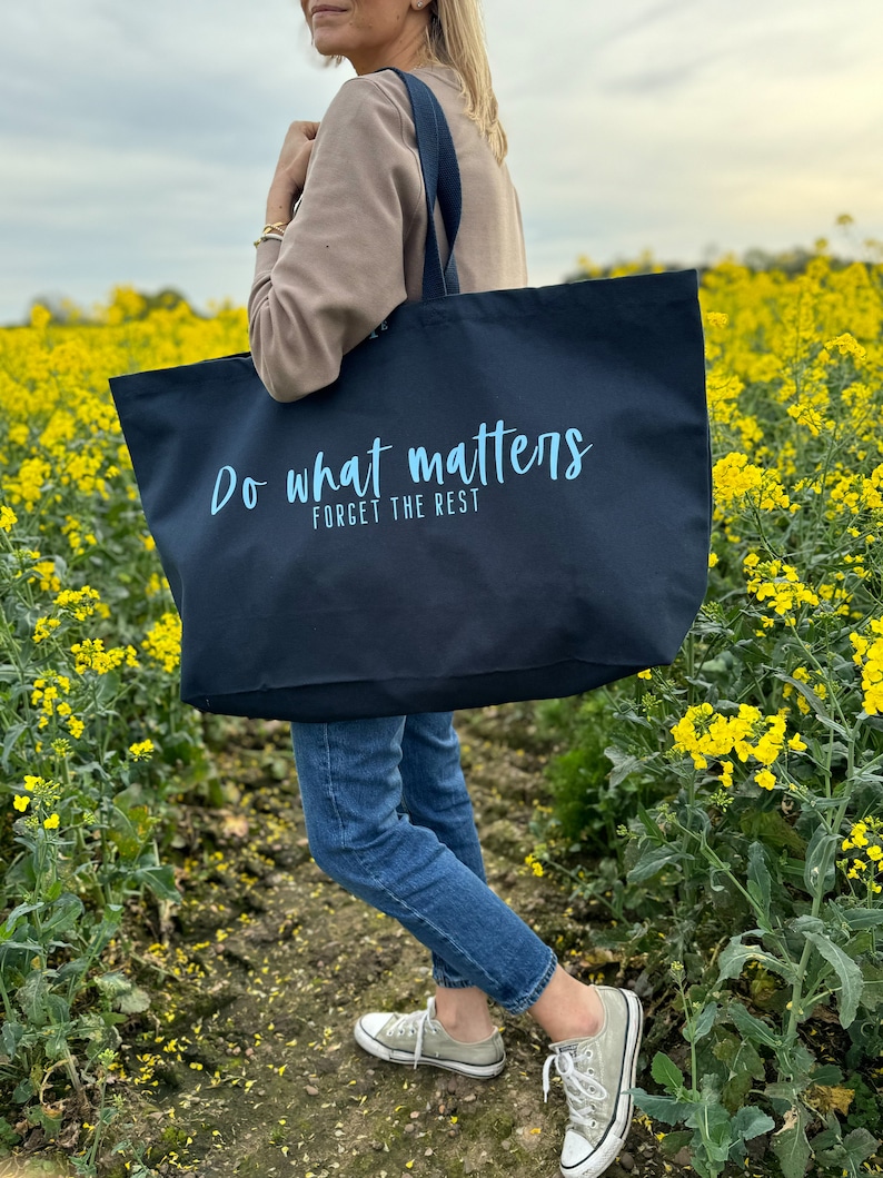 Pode incluir: Uma bolsa tote preta com o texto "Do what matters, forget the rest" em letras azuis claras. A bolsa est&aacute; sendo segurada por uma pessoa que est&aacute; vestindo jeans azul e um su&eacute;ter marrom.