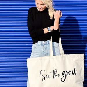 Pode incluir: Uma bolsa tote de lona branca com texto preto que diz "See the good".