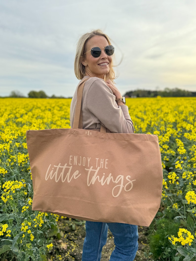 Pode incluir: Uma bolsa tote de lona marrom com o texto "Enjoy the little things" em branco. A bolsa est&aacute; sendo segurada por uma pessoa usando &oacute;culos de sol e um su&eacute;ter marrom em um campo de flores amarelas.