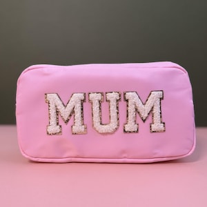 Pode incluir: Bolsa rosa com zíper com a palavra "MUM" em letras brancas com detalhes de glitter dourado.