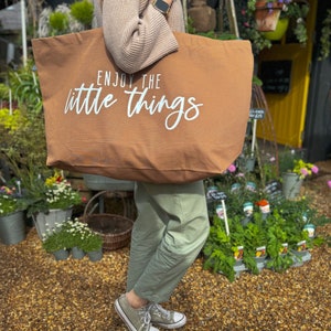 Pode incluir: Uma bolsa tote de lona marrom com o texto "Enjoy the little things" em letras brancas. A bolsa est&aacute; sendo segurada por uma pessoa vestindo um su&eacute;ter marrom claro e cal&ccedil;as verdes.