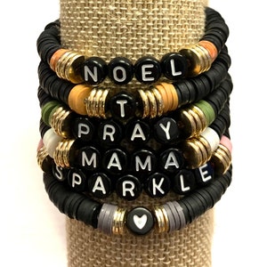 May include: A stack of black beaded bracelets with gold accents and white lettering spelling out the words "NOEL", "T", "PRAY", "MAMA", and "SPARKLE". A small heart charm is at the bottom of the stack.
