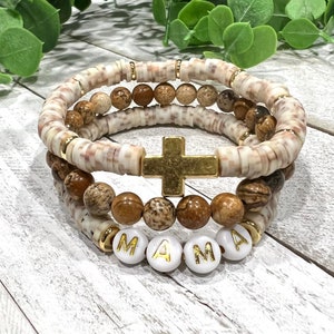 May include: A set of three bracelets featuring brown and white beads, a gold cross charm, and white letter beads spelling "MAMA".