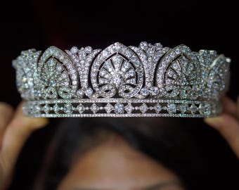 May include: A silver tiara with intricate detailing, featuring a series of arched designs and sparkling gemstones. The crown is held up against a dark background, showcasing its elegant design.