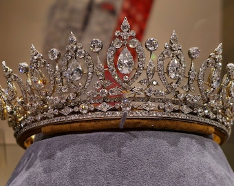 May include: A detailed view of a diamond and gold crown. The crown features numerous clear, sparkling diamonds of various shapes and sizes, set in a gold filigree design. The crown is displayed on a gray velvet surface.