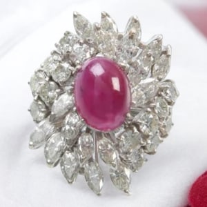 Könnte beinhalten: Ein auffälliger Ring mit einem großen, ovalen, tiefroten Edelstein im Zentrum, umgeben von einer Ansammlung kleinerer, klarer, navetteförmiger Steine. Die Fassung des Rings ist silberfarben und bildet einen Kontrast zum roten Edelstein.