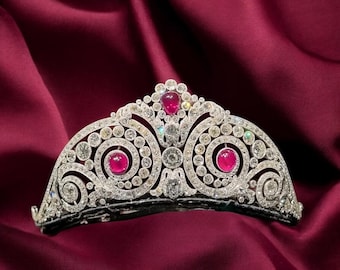May include: A silver tiara with a repeating swirl design, encrusted with numerous small diamonds and three large, oval-cut red gemstones. The tiara sits on a deep red satin background.