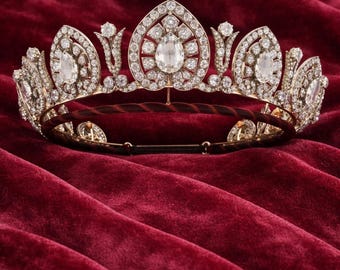 May include: A gold-toned tiara with a central heart-shaped design, adorned with numerous clear gemstones. The tiara's intricate details include teardrop-shaped accents and a dark brown band, set against a deep red velvet backdrop.