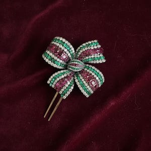 May include: A jeweled bow hair clip featuring a design of alternating green and ruby-colored stones, accented with clear crystals. The bow is set on a gold-toned metal base with two prongs.