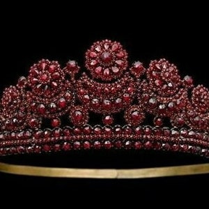Peut inclure: Une couronne en or incrustée de centaines de pierres précieuses rouge foncé. La couronne a un groupe central de pierres plus grand et deux groupes plus petits de chaque côté.