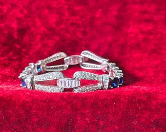 May include: A silver-toned bracelet featuring a series of interconnected links. The design incorporates alternating rectangular blue stones and clear, sparkling stones set into the metal frame, creating a continuous, repeating geometric pattern.