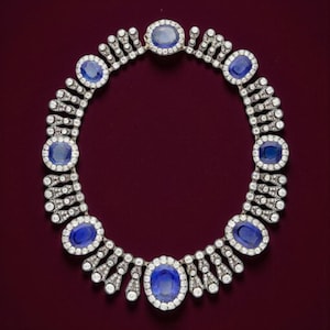 May include: A stunning necklace featuring large oval blue gemstones set in a silver or white metal. The necklace is adorned with numerous small, clear gemstones, creating a sparkling, elegant design. The necklace is displayed against a deep red background.
