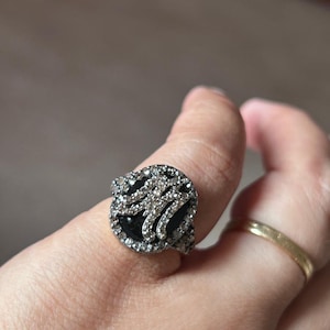 May include: A ring featuring a circular black stone centerpiece surrounded by a border of small clear stones. The letter M is prominently displayed in the center, encrusted with small clear stones. The band is textured with a metallic finish.