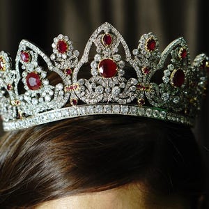 May include: A silver tiara with red and clear gemstones. The crown features a series of arches, each adorned with a large, round ruby. Smaller rubies and clear crystals are set throughout the design, creating a sparkling effect. The tiara sits atop dark brown hair.