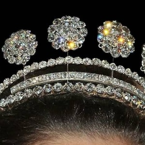 Puede incluir: Una tiara de diamantes brillantes con una banda de plata y cinco grandes racimos de diamantes. La tiara está colocada sobre un fondo oscuro.
