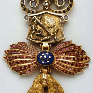 Op de afbeelding: Een gouden hanger met een blauwe en gouden emaille strik, omringd door rode edelstenen. De hanger heeft een gouden schild met een scène van een man te paard, en een kroon met blauwe en witte edelstenen.