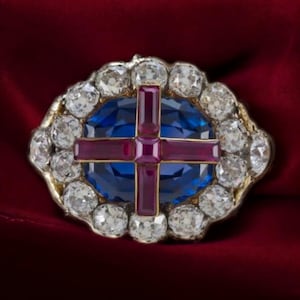Puede incluir: Un broche de oro y plata con un zafiro azul rodeado por un halo de diamantes y una cruz roja.