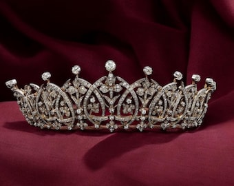 May include: An ornate, antique tiara with a silver-colored metal frame. The tiara is encrusted with numerous clear gemstones, and features a repeating floral and arched design. The background is a deep red fabric.