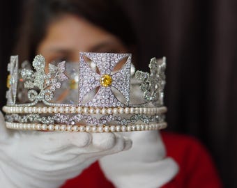 May include: A silver crown with a large yellow gemstone in the center, surrounded by smaller white gemstones. The crown is held in a gloved hand.