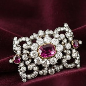 May include: An ornate brooch featuring a large, oval, magenta-colored gemstone at its center, surrounded by a halo of smaller, clear gemstones. Additional magenta gemstones accent the sides. The brooch is set in a silver-toned metal with intricate detailing.