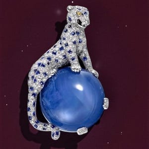 Könnte beinhalten: Eine mit Juwelen besetzte Leopardenfigur mit silbernem Körper, blauen Saphirflecken und gelben Augen, die auf einem großen, runden, blauen Edelstein sitzt. Die Figur ist vor einem dunkelroten Hintergrund platziert.