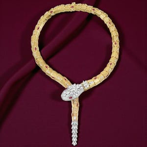 Op de afbeelding: Een gouden slangenketting met rode en witte accenten. De ketting heeft een slangenkop met een rode edelsteen oog en een diamantachtig patroon. Het lichaam van de slang is gesegmenteerd met rode en witte details.