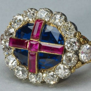 May include: A gold ring featuring a large blue sapphire surrounded by a halo of diamonds. The sapphire is set with a red ruby cross.