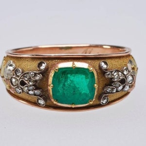 Puede incluir: Un anillo de oro con una gran esmeralda rodeada de diamantes más pequeños. El anillo está engastado en una banda de oro con una superficie texturizada.