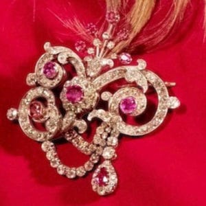 Op de afbeelding: Een sierlijke, zilverkleurige broche met een wervelend ontwerp, ingelegd met heldere kristallen en levendige roze edelstenen. Een druppelvormige roze edelsteen hangt aan de onderkant. De broche staat tegen een rode achtergrond.