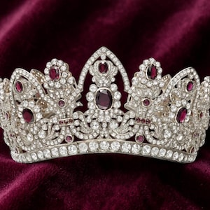 May include: A silver-toned tiara featuring a central pointed arch and multiple smaller arches, all encrusted with numerous clear white crystals and accented with deep red oval-shaped stones arranged in a symmetrical, intricate floral pattern.