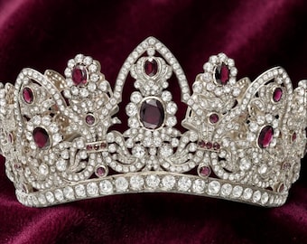 Op de afbeelding: Een zilverkleurige tiara met een centrale puntboog en meerdere kleinere bogen, ingelegd met talloze heldere witte kristallen en geaccentueerd met donkerrode ovale stenen in een symmetrisch, ingewikkeld bloemmotief.