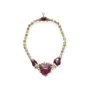 Può includere: Una collana d'oro con un ciondolo di pietra rossa e pietre rosse più piccole lungo la catena. La collana è decorata con piccoli diamanti.