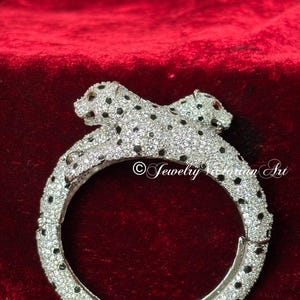 Pode incluir: Uma pulseira rígida articulada de tom prateado com duas figuras de leopardo que se encontram no topo. Toda a superfície é incrustada com pedras pavé transparentes e pequenas pedras pretas que formam manchas. Texto Jewelry Victorian Art visível.