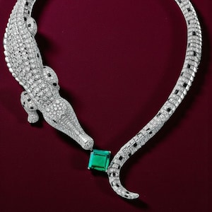 Op de afbeelding: Een luxe ketting met een krokodillenontwerp, gemaakt van diamanten en een smaragd. Het lichaam van de krokodil is gedetailleerd met diamanten en zwarte accenten. De ketting heeft een grote, vierkant geslepen smaragd in het midden.