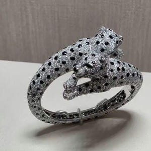 Pode incluir: Uma pulseira rígida articulada de tom prateado em forma de leopardo enrolado. Toda a superfície está incrustada com pequenas pedras pavé transparentes e acentuada com pedras pretas dispostas num padrão de manchas pelo corpo e cabeça do felino.