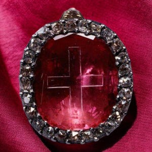 May include: A red gemstone pendant with a cross etched into it. The gemstone is surrounded by a silver setting with small diamonds.